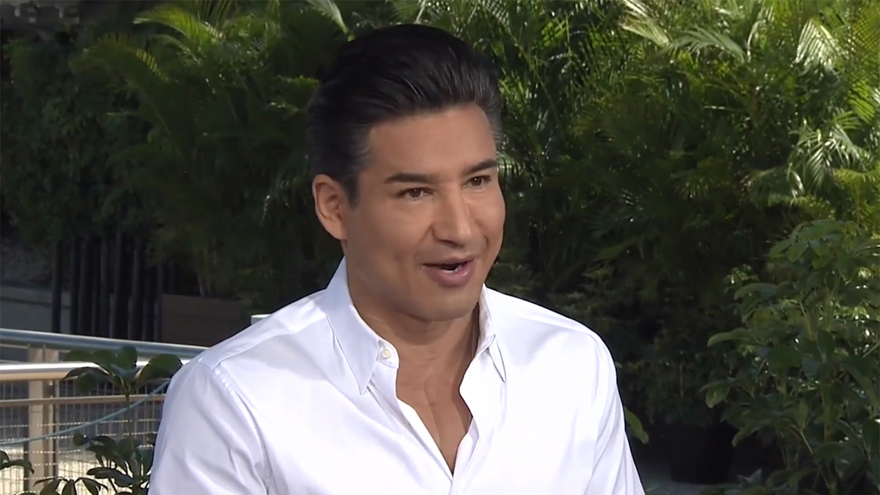 Mario Lopez filming at a desk set up near the Jurassic area of Universal Orlando. He is in a white shirt partially unbuttoned.