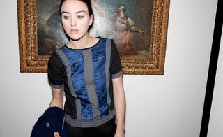 Female model wearing a black blue and grey top, black pants, navy blue coat draped over her arm, white wall background, painting in bronze colour frame on the wall