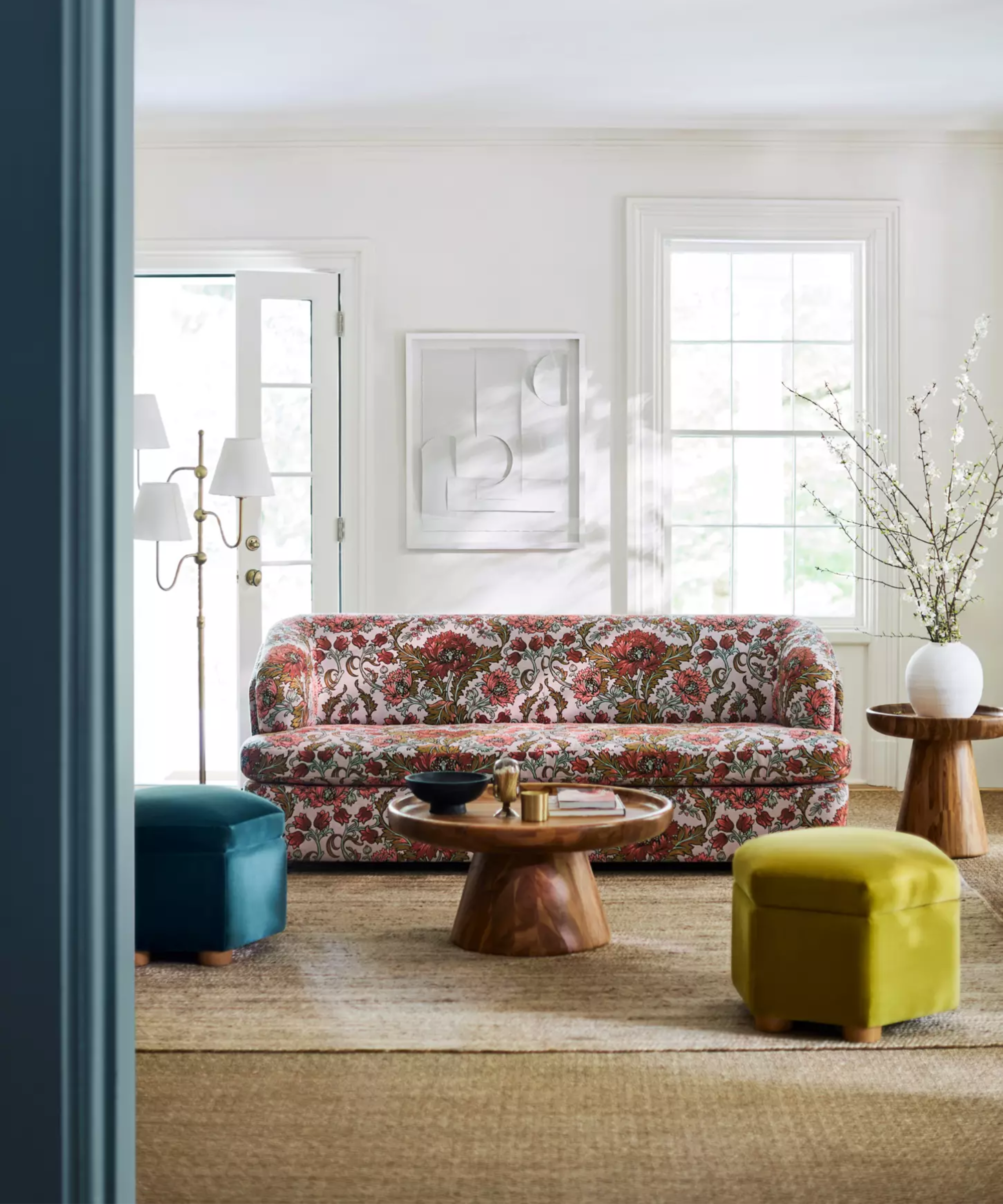 Floral jacquard sleeper sofa styled in a bright, white artistic living room featuring a three-shade floor lamp, jewel-toned ottomans, and wooden side and coffee tables.