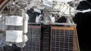two astronauts wearing white spacesuits work outside of a space station "above" a pair of solar array panels