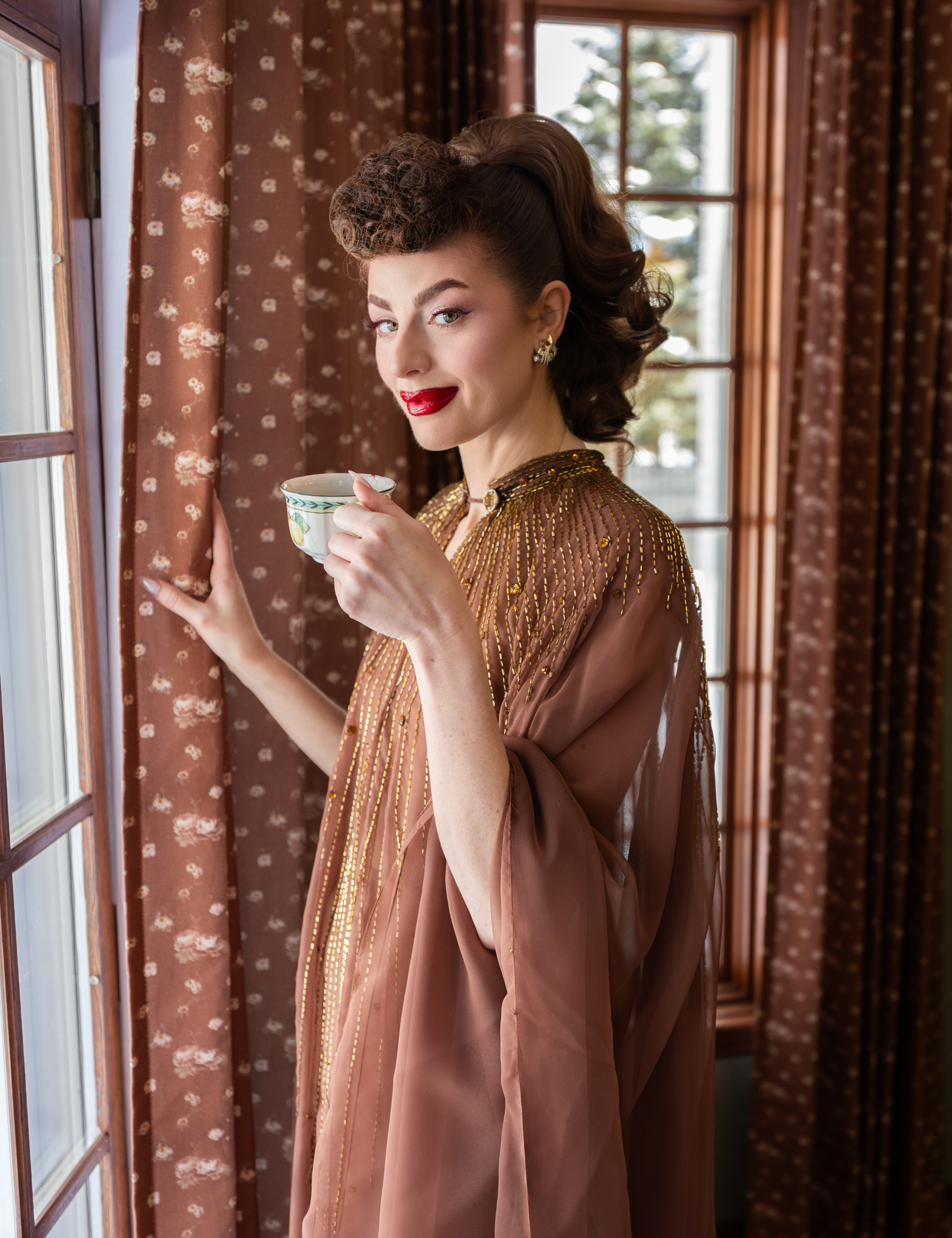 Ms. Maverick Muse poses in a wood-clad dining room area featuring large windowpanes and brown botanically patterned curtains