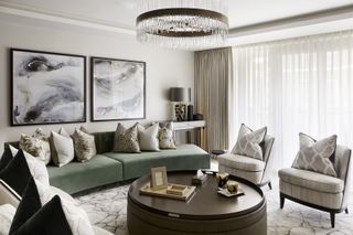 neutral living room with green curvy couch large square artwork on walls, curvy armchairs, round coffee table, wood top, chandelier, drapes, patterned cushions