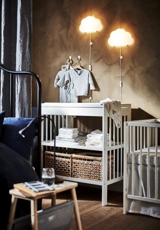 nursery with white crib, white changing unit with open storage underneath, wooden floor, gray drapes, cloud lights, textured walls
