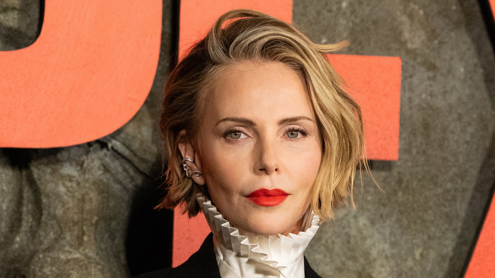 Image of Charlize Theron with blonde chin-length bob, wearing red lipstick and a white high-neck pleated shirt