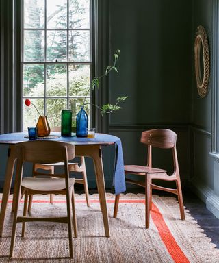 Dining room with dark blue painted walls, rounded mirror with textured, woven frame, dark wood, round dining table with matching dining chairs, blue table throw, textured, natural rug with orange stripe and scalloped edging, black wooden flooring, decorative vases and glass ornaments on table