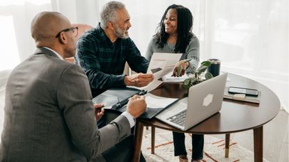 A couple sit at a round table looking at paperwork with a financial adviser who has his laptop open.