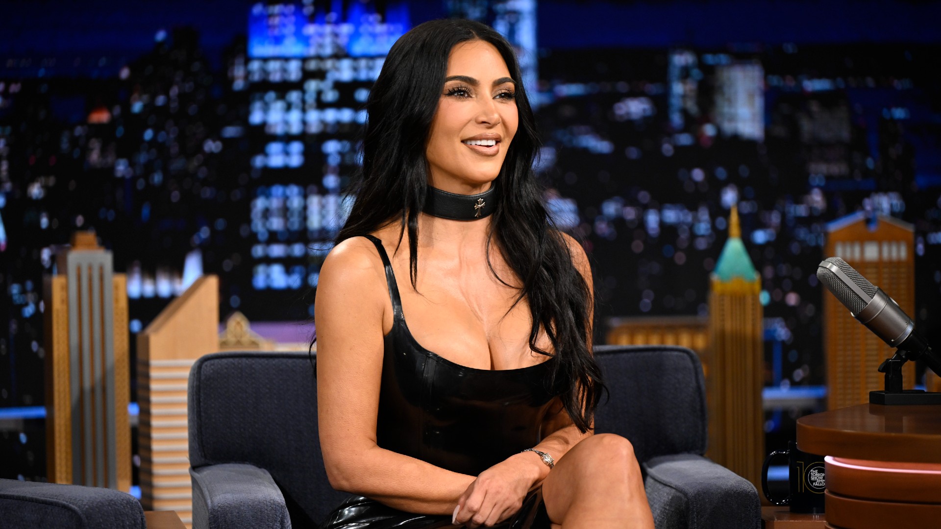 a woman with long, dark hair and a black, strappy dress sitting on a talk show set in front of a live studio audience