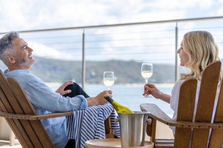 Mature couple drinking wine out on the deck. They are happy and smiling sitting in deck chairs. The sea is in the background.