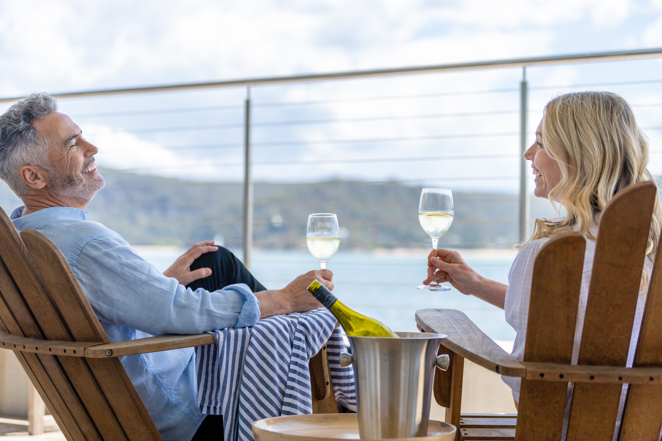 Mature couple drinking wine out on the deck. They are happy and smiling sitting in deck chairs. The sea is in the background.
