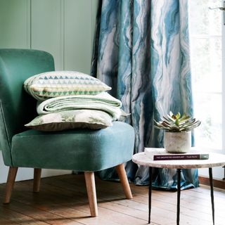 forest green armchair with pillows on wooden flooring against a blue and cream patterned curtain