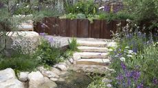 Flower beds surround a stone pathway, steps and a pond in a show garden