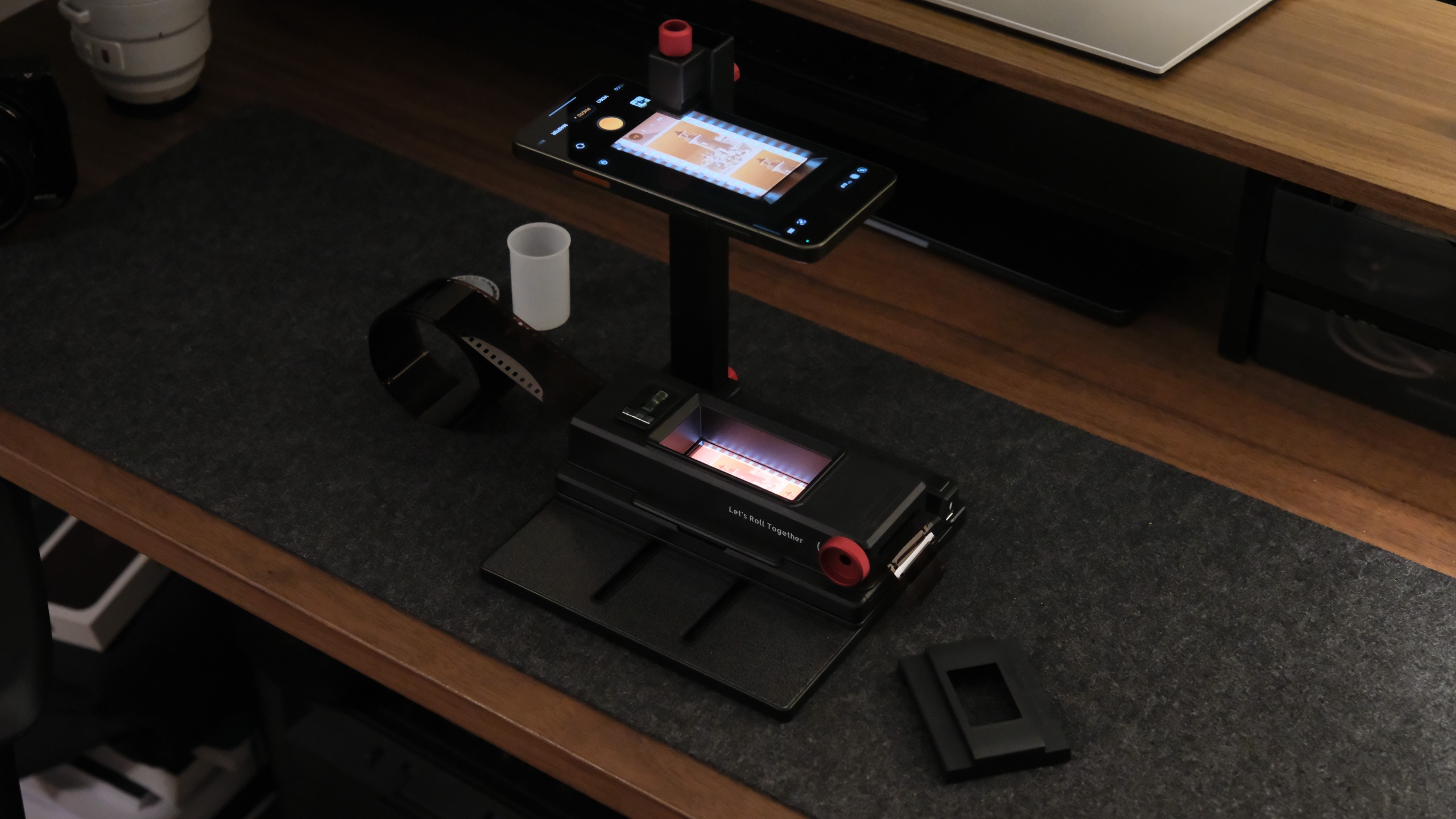 A complete film scanning workstation on a wooden desk, showing a smartphone attached to a vertical copy stand capturing an illuminated strip of 35mm film on a grey mat.