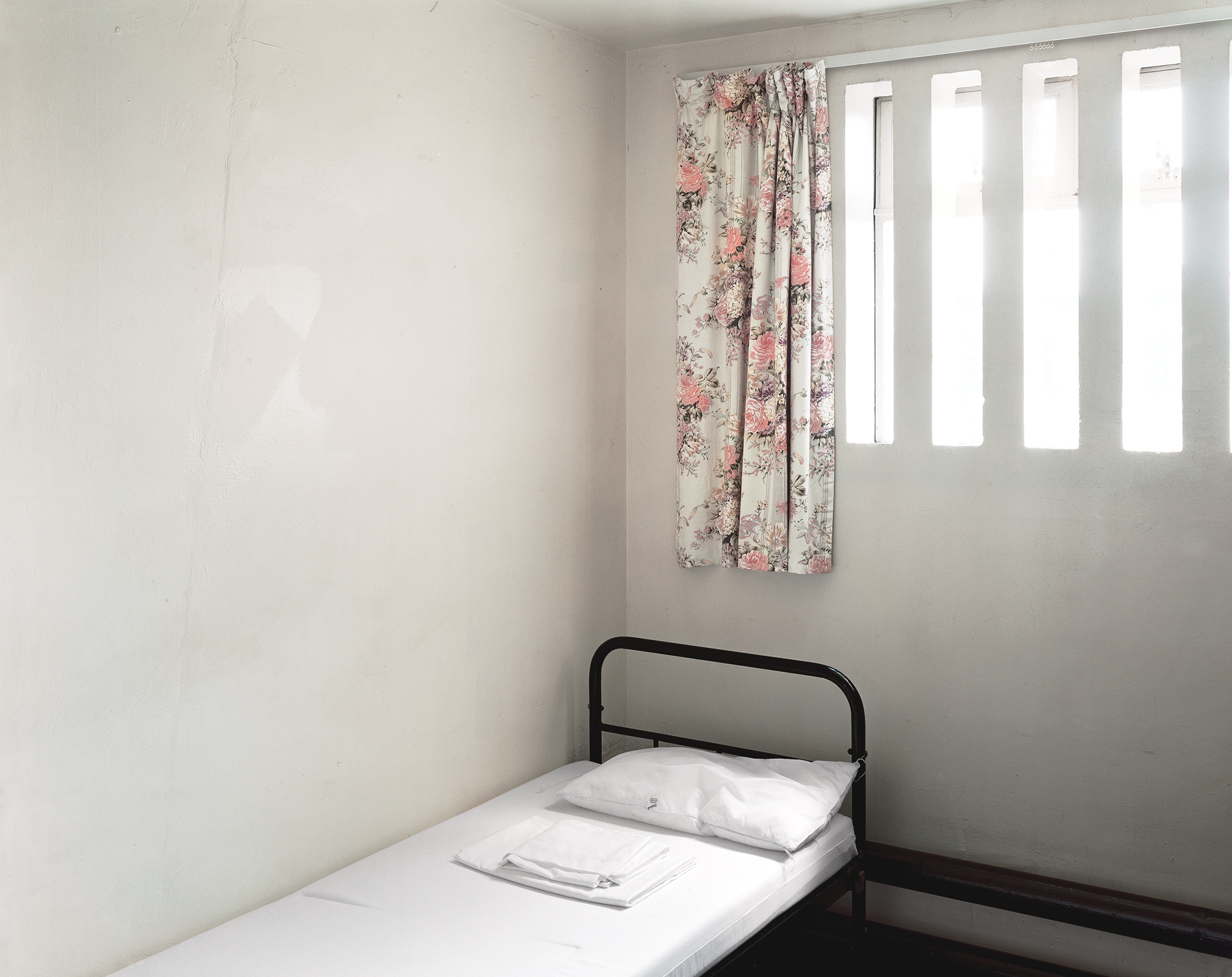 Colour photograph of a stark, whitewashed prison cell with a single made bed and a barred window softened by a floral curtain.