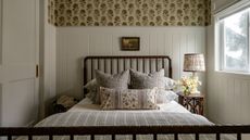 A cozy guest bedroom with warm white paneling, ditsy floral wallpaper on the upper walls, and built-in wardrobes. A dark wood spindle bed frame with neutral patterned bedding, and a patterned table lamp. 