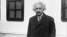 A black and white photo of a white haired man with a dark moustache and a dark blazer with tie.