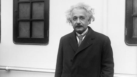 A black and white photo of a white haired man with a dark moustache and a dark blazer with tie.