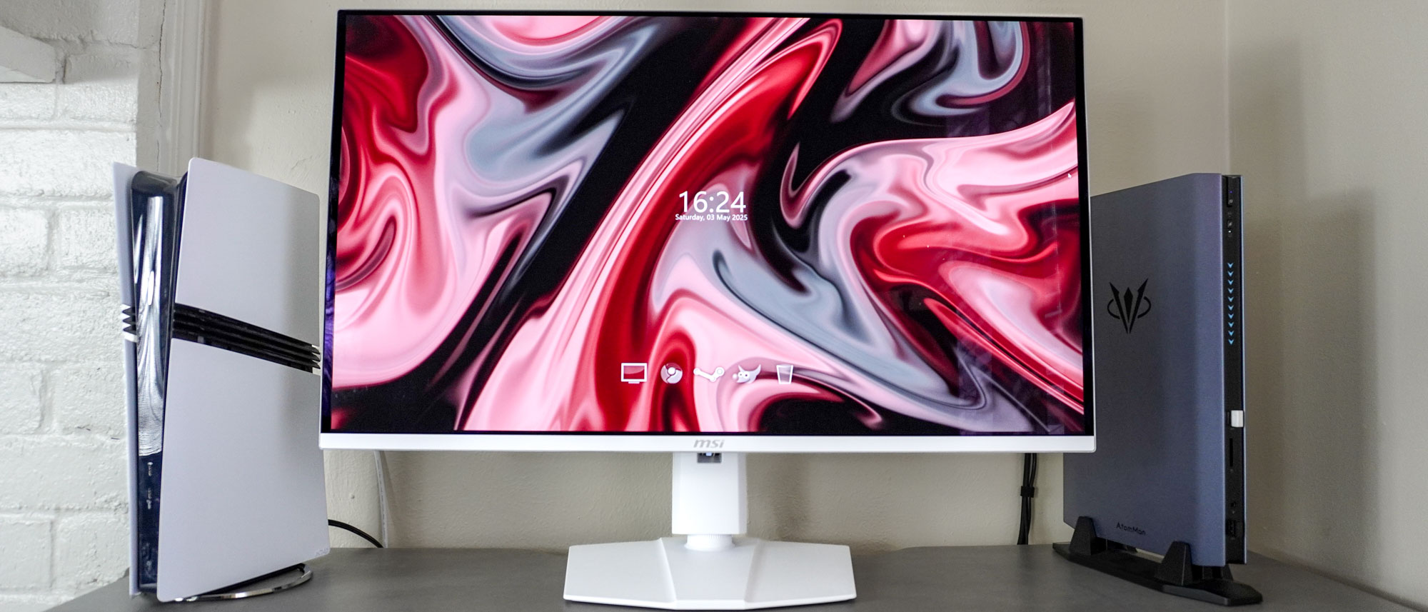 The MSI MPG 321URXW QD-OLED monitor on a desk next to a PS5 Pro and the Atomman G7 Ti mini gaming PC