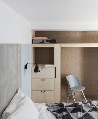 A room in the Leman Locke hotel. To the far wall, we see an open closet space, with a gray chair. Closer to us, we see a part of the bed, with a gray and black blanket with a geometrical pattern.