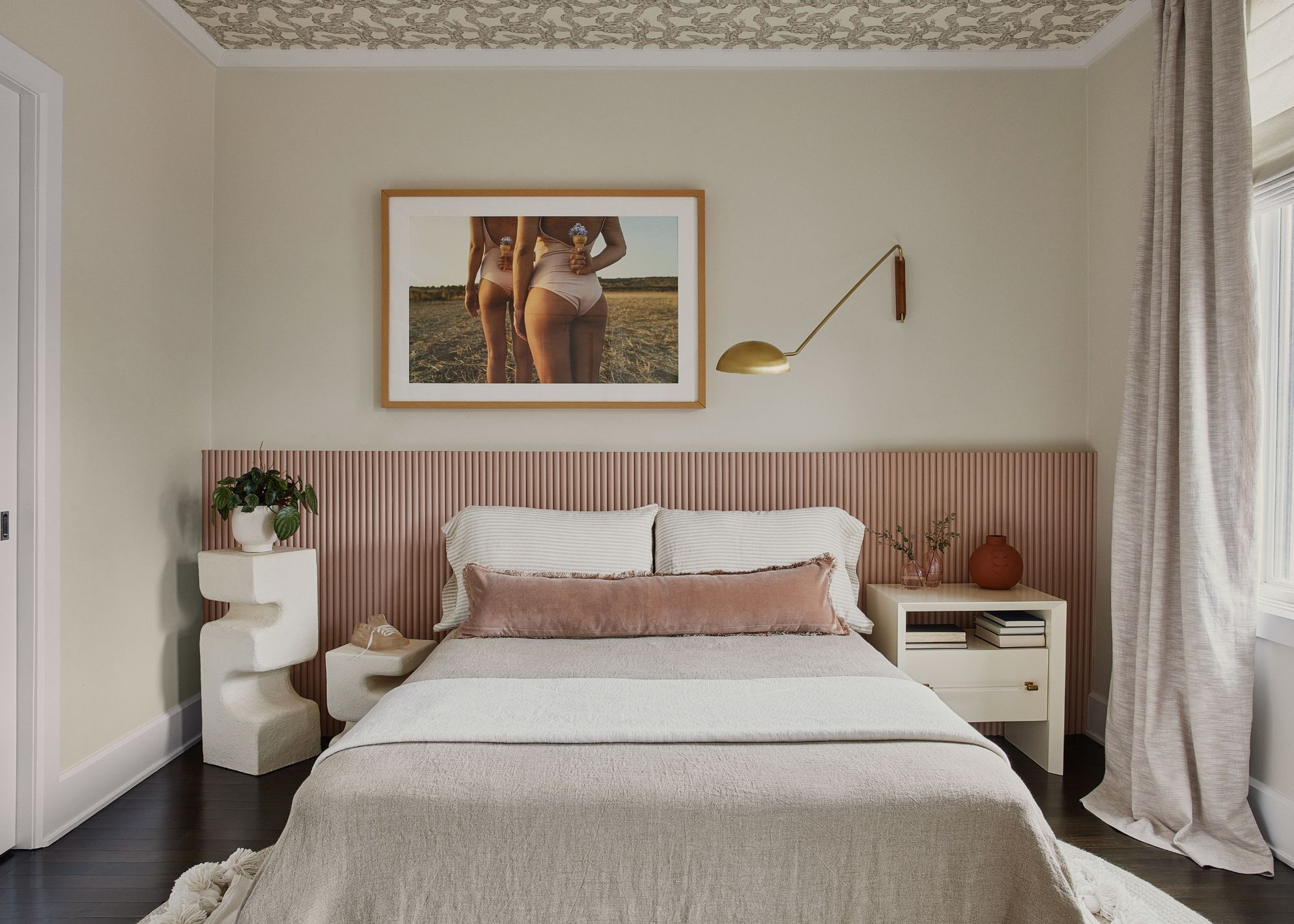 A bedroom with a bed with a pink headboard, a designer-side table and a ceiling wallpaper