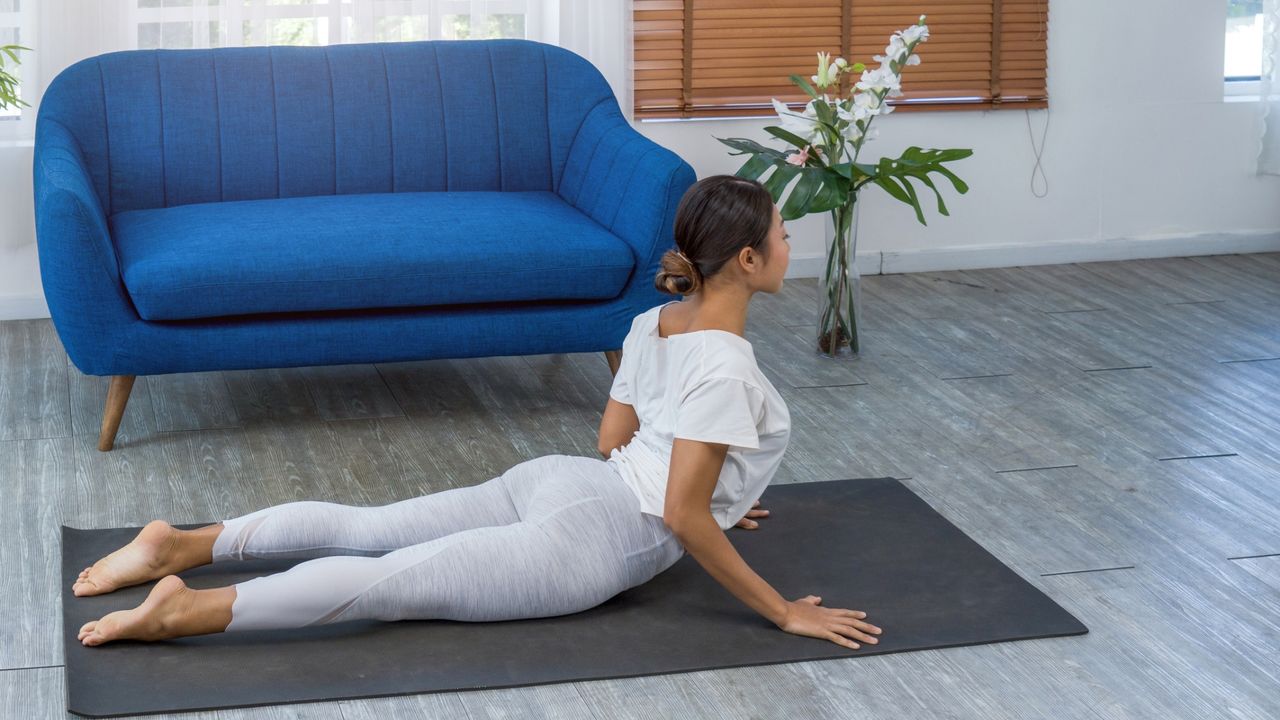 woman doing a cobra back stretch pose on a grey yoga mat in a living room setting with a blue sofa behind her