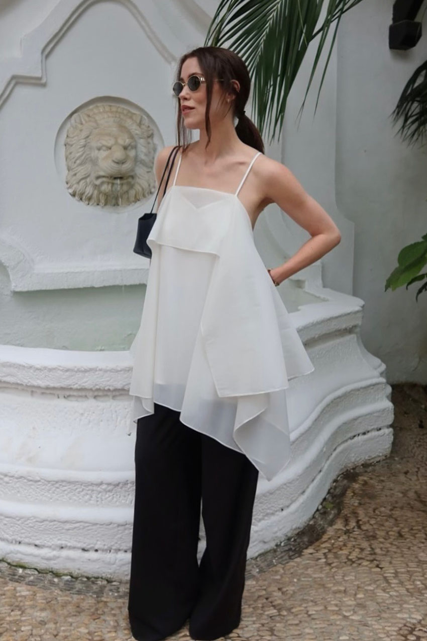 A woman wearing a white sleeveless top, black pants, a black shoulder bag, and black sunglasses.