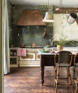 Green tiled backsplash with wooden parquet flooring, wooden kitchen table, copper oven hood, copper pans and cream cabinetry