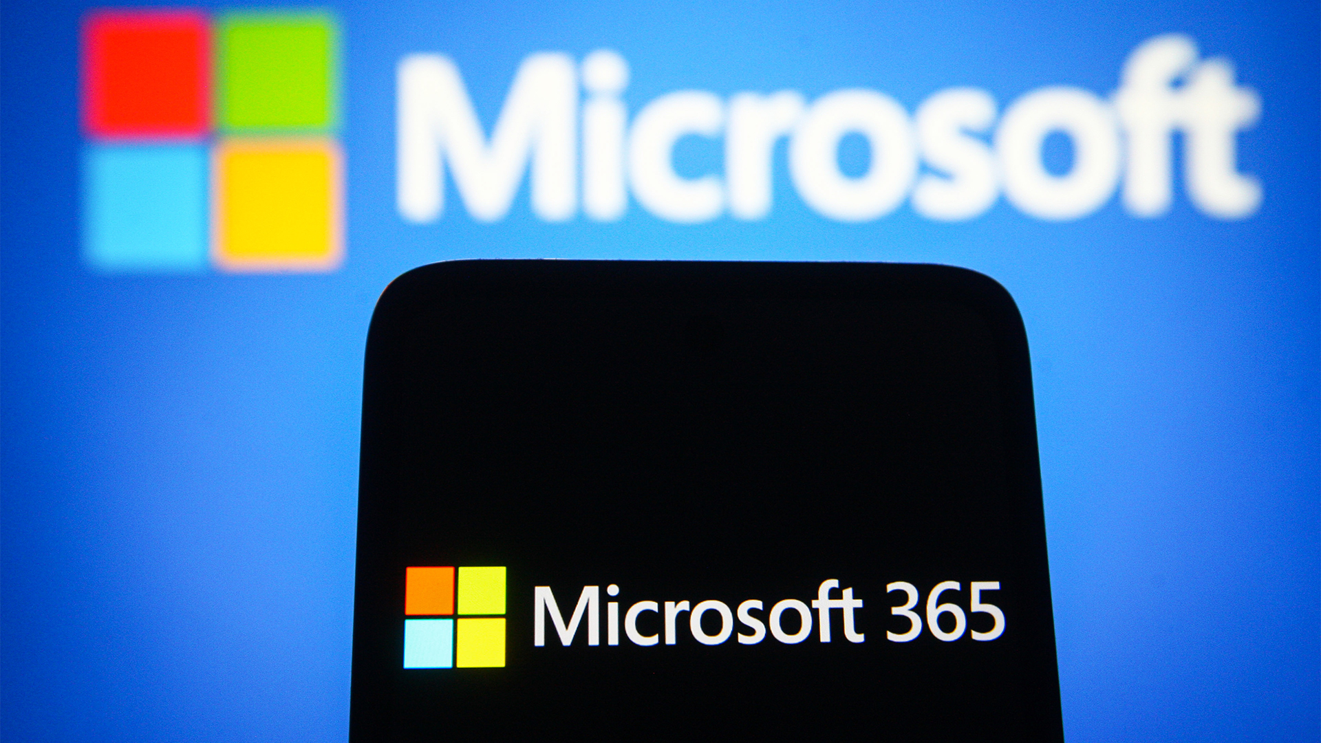Microsoft 365 logo and branding pictured on a smartphone screen with Microsoft logo and branding pictured in background.