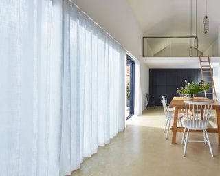 Open plan kitchen-diner with large floor to ceiling windows with white sheer curtains, cream flooring, wooden dining table, wooden ladder up to mezzanine