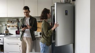Woman opening a silver refrigerator with a man stood next to her on an ipad. 