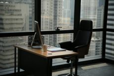 An empty office desk and seat in front of windows in a city at dusk.