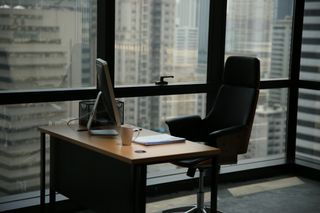 An empty office desk and seat in front of windows in a city at dusk.