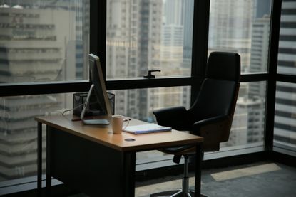 An empty office desk and seat in front of windows in a city at dusk.