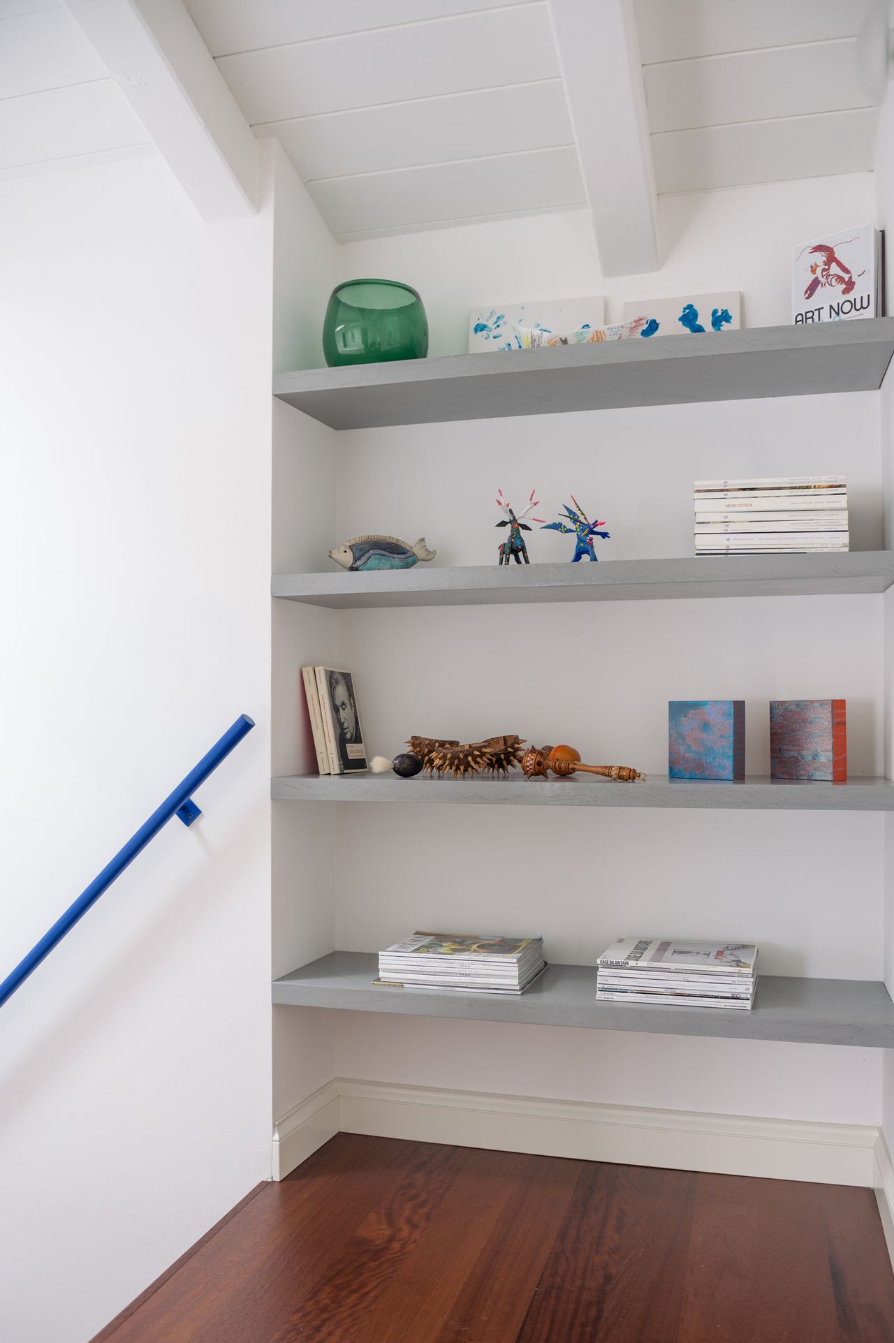 Image of a white hallway and staircase with built-in floating shelves and hardwood floors. There is an electric blue handrail in view.