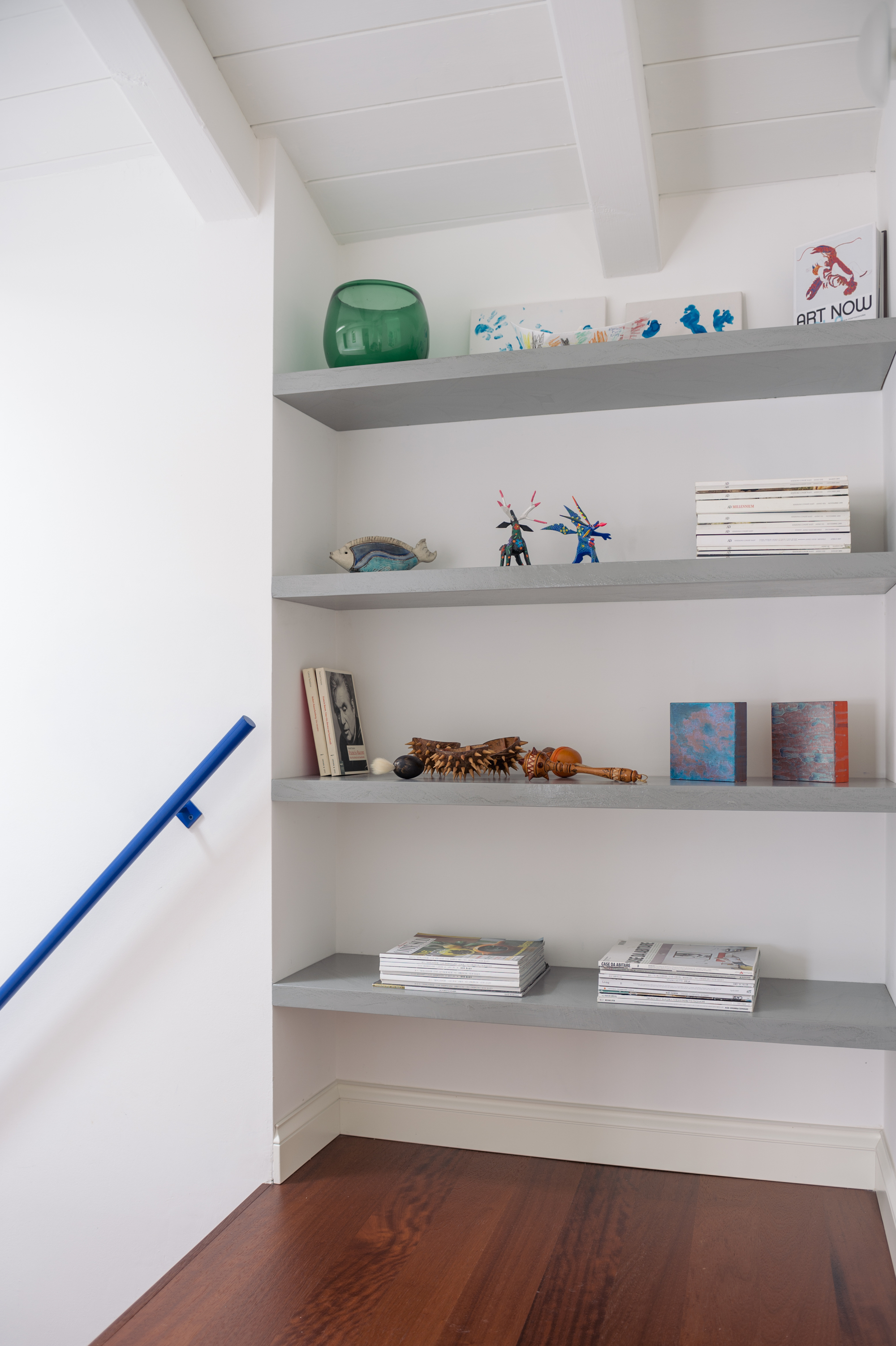 Image of a white hallway and staircase with built-in floating shelves and hardwood floors. There is an electric blue handrail in view.