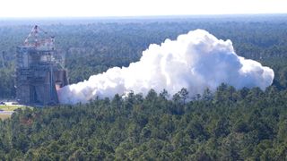 An industrial construct stands amidst a green forest with smoke billowing out into a plumeA full duration test of the RS-25 certification engine was conducted at NASA's Stennis Space Center on October 17, 2023.
