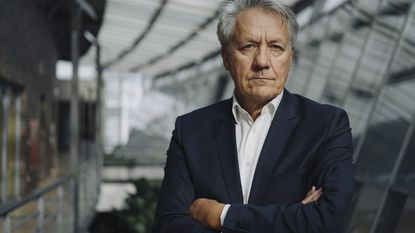 An older man stands in an office with his arms crossed and a stern look on his face.