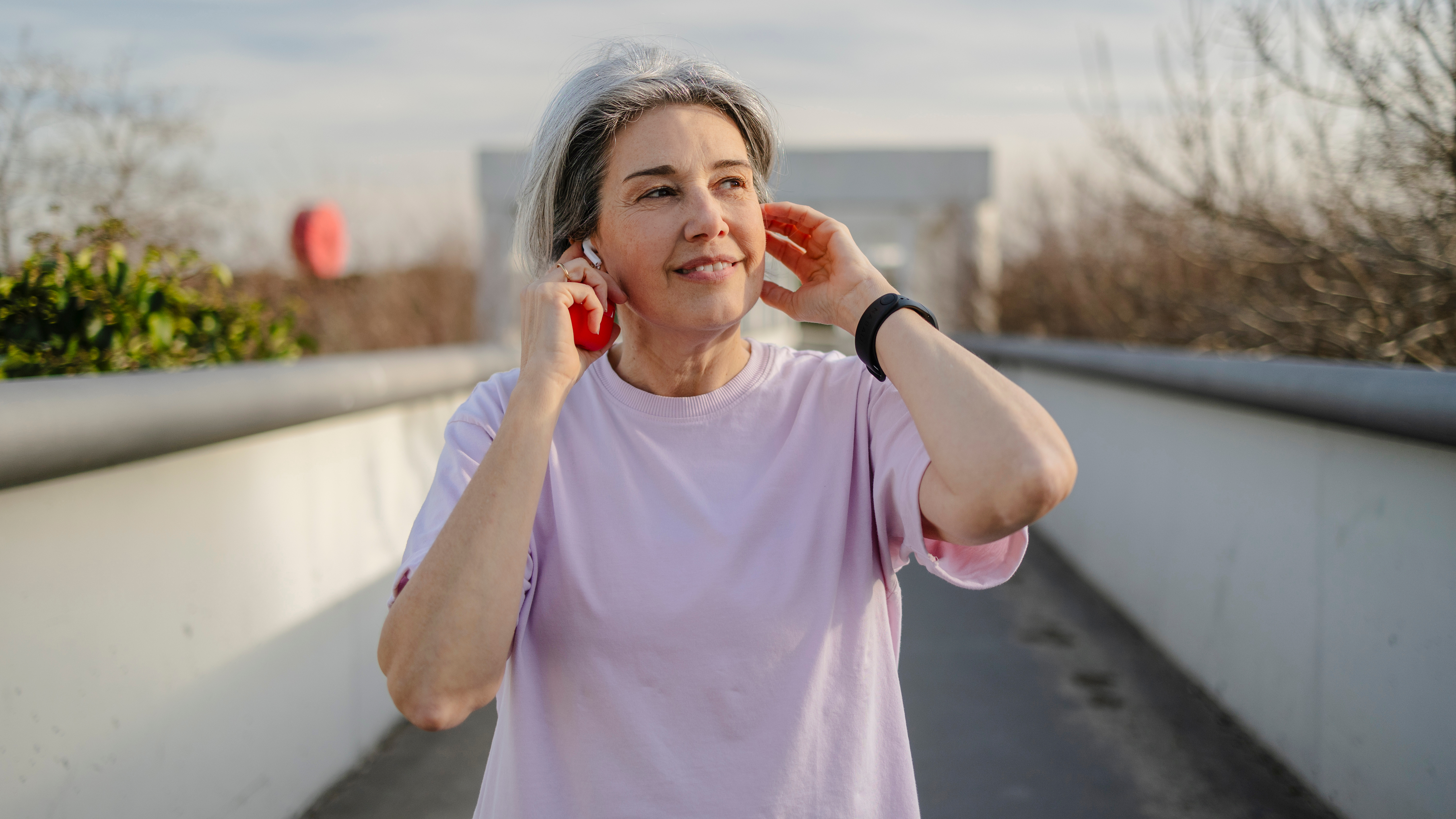 A woman walks along an overpass, putting in some earbuds and holding an apple in her right hand. 