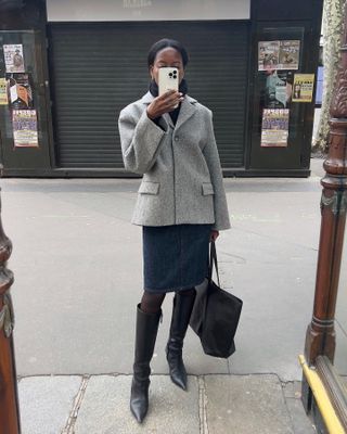 Sylvie wears a gray blazer, denim skirt, black tights, black knee-high boots, and a black tote bag.