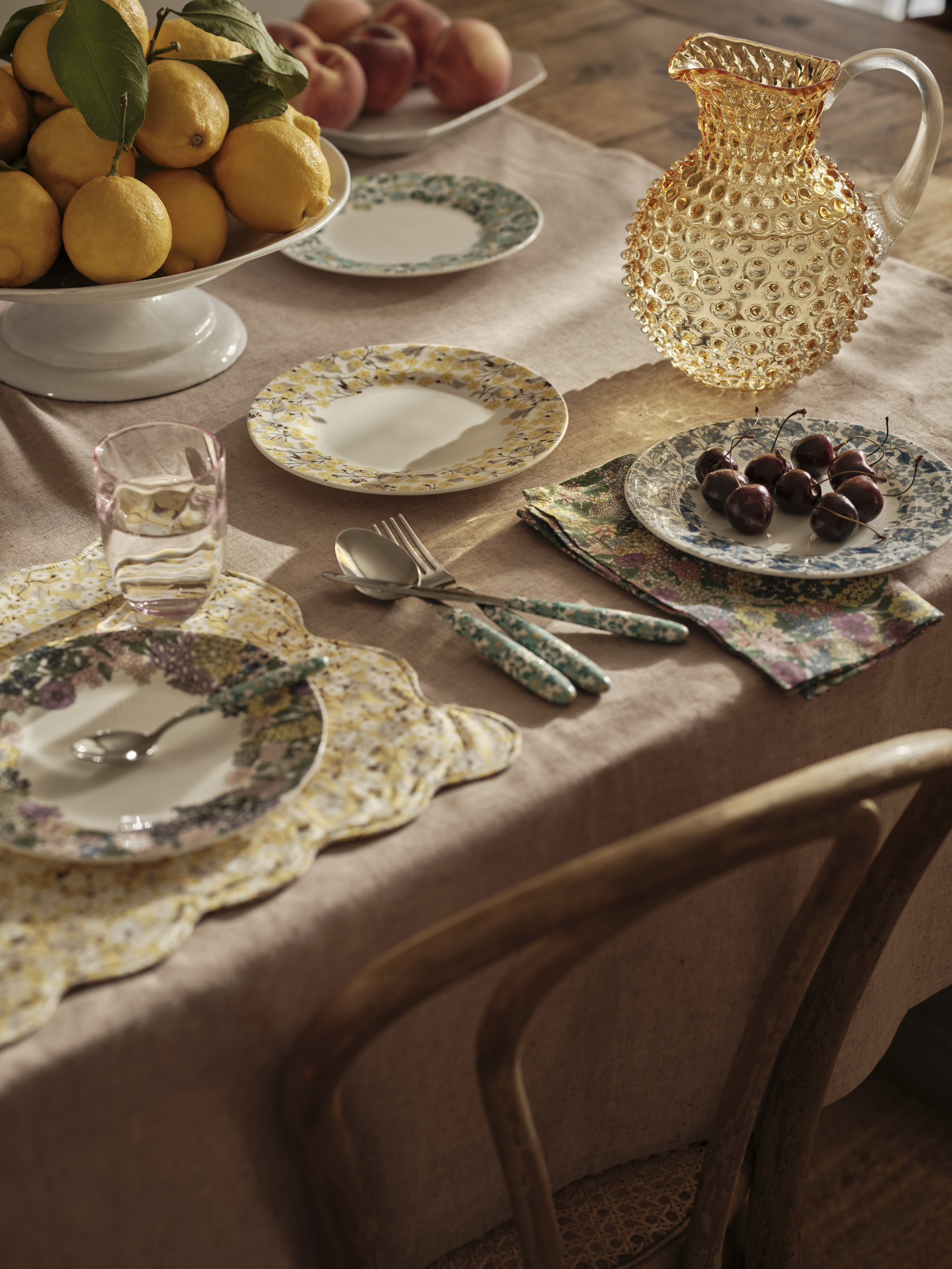 a dining table with a light pink tablecloth, floral plates and cutlery, and a bowl of citrus