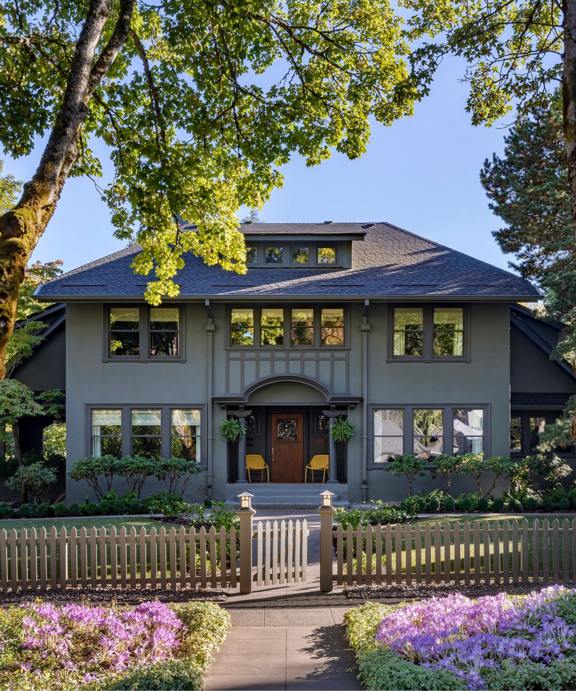 A home exterior with muted green siding, dark green trim, a dark gray roof, and a brown front door. A gated front garden and purple flowers in the foreground.