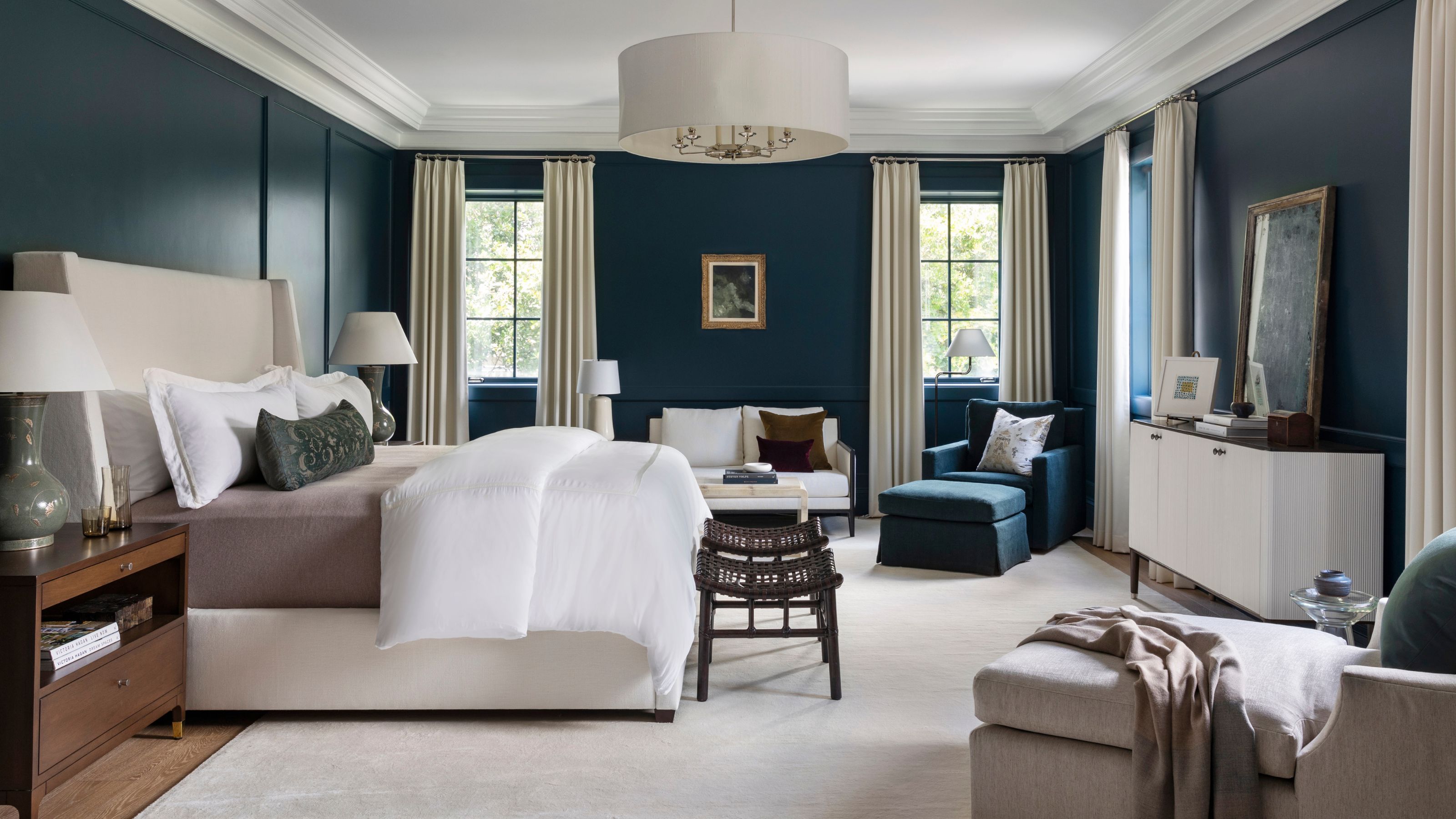 a navy primary suite with neutral bed, a small sitting area with a white couch, navy armchair and dark wooden nightstand