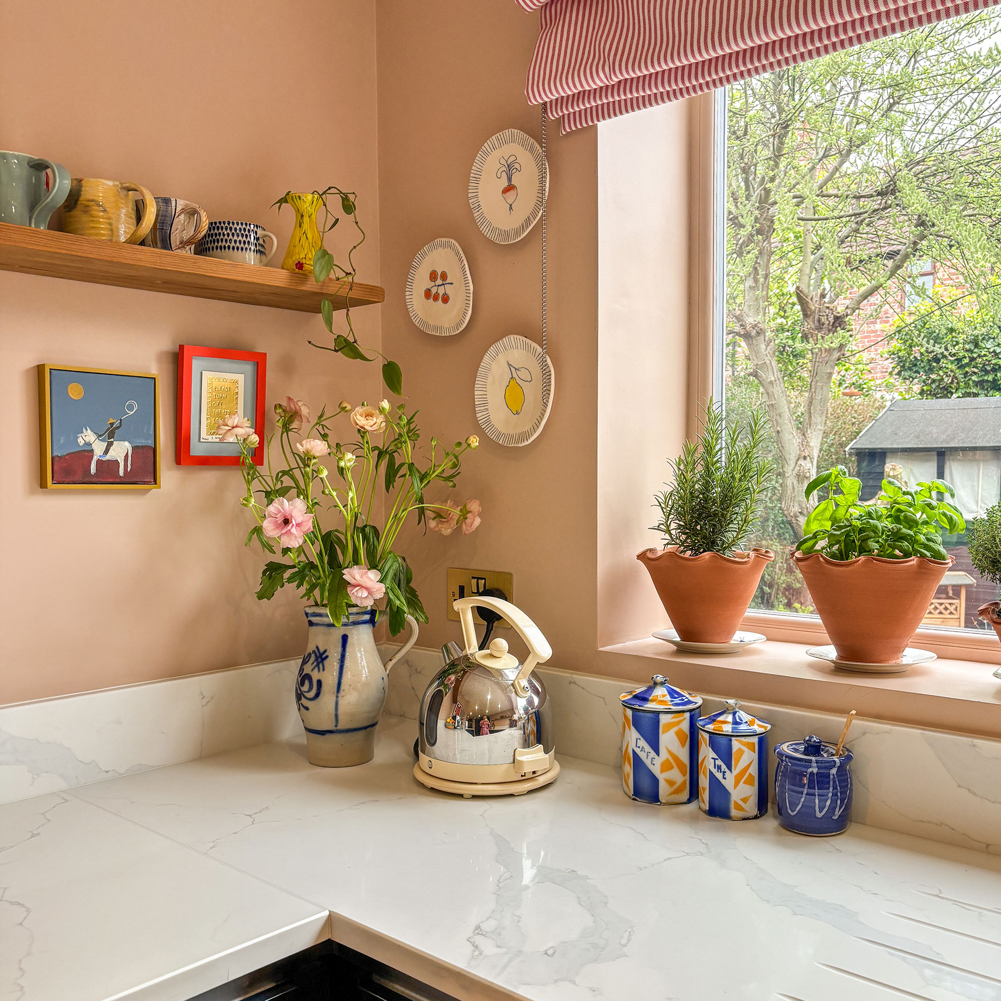 Corner of peach kitchen with quartz worktops, pots of herbs on windowsill and display plates on wall