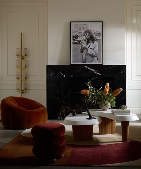 Close up of furniture in front of a black marble fireplace, mantel decorated with artwork, sculptural metallic wall light to the left hand side, geometric rug underneath furniture