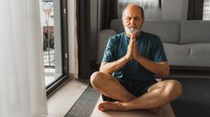 An older man sits on his living room floor looking very relaxed while he does yoga.