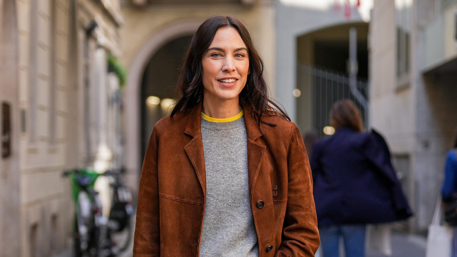 Alexa Chung wearing a Miu Miu brown suede jacket with a grey sweater over a yellow T-shirt, baggy jeans, and ballet flats in Milan