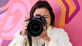 A photographer holding a Nikon camera in front of their face, standing in front of a pink-and-yellow background