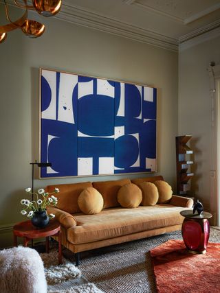 A blue painting hanging above an orange-toned velvet sofa with red accent details around