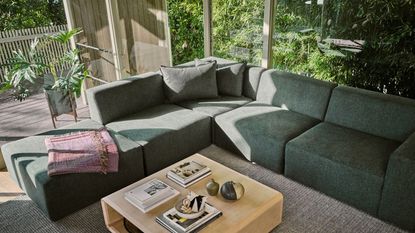 Best couch. Green corner sofa in bright living room with large floor to ceiling windows, lots of greenery and plants in background, rug, light wooden coffee table