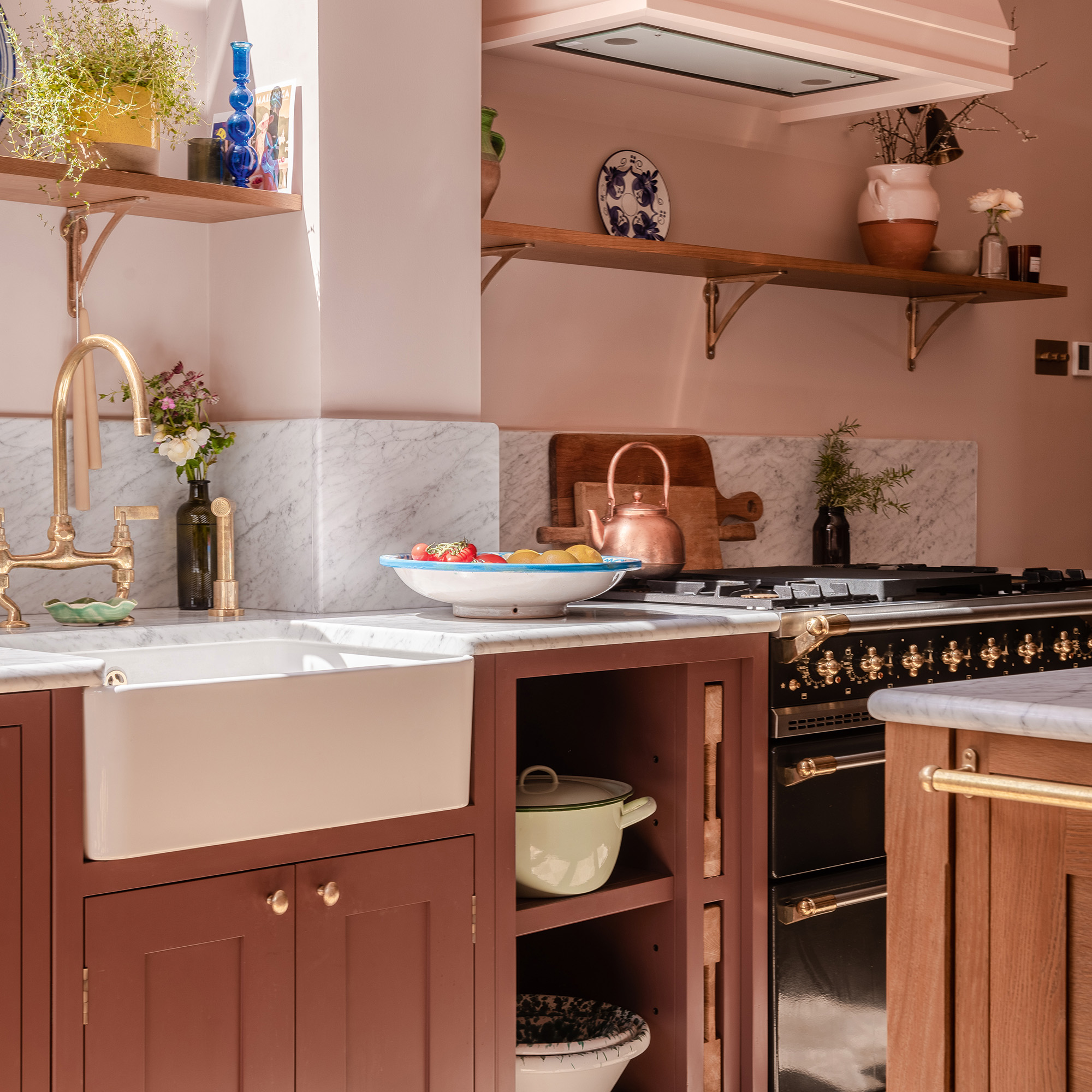 close up of butler sink and brass taps next to black range cooker in kitchen with pink walls and pink painted extractor fan and purple cabinetry with marble worktop and splashback as well as an open shelf herringbone house