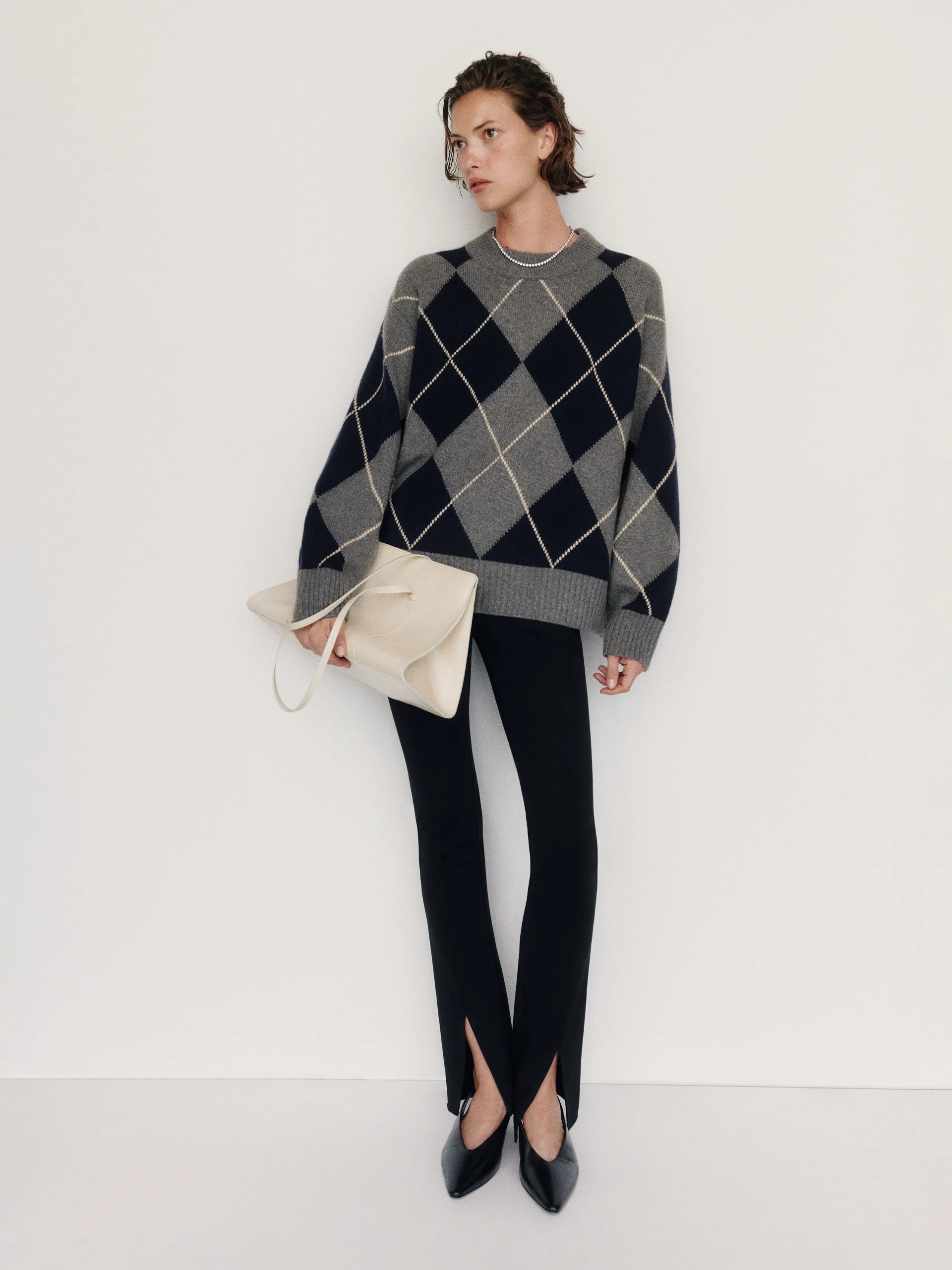 An image. ofa model wearing a grey and black relaxed argyle jumper with black trousers with split hems